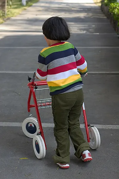 Kind mit buntem Pullover nutzt einen Anterior-Walker zur Mobilitätsunterstützung beim Gehen auf einem Bürgersteig im Freien.
