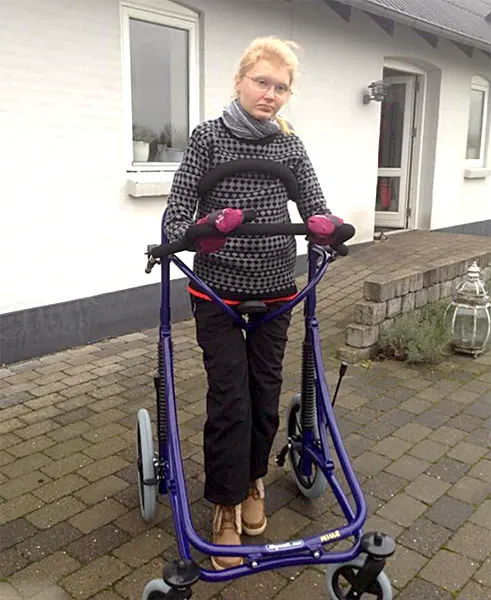 Junge Frau (Mille) mit Brille und Handschuhen nutzt einen violetten VELA Meywalk 2000 Gehwagen draußen auf gepflastertem Weg vor einem Haus.