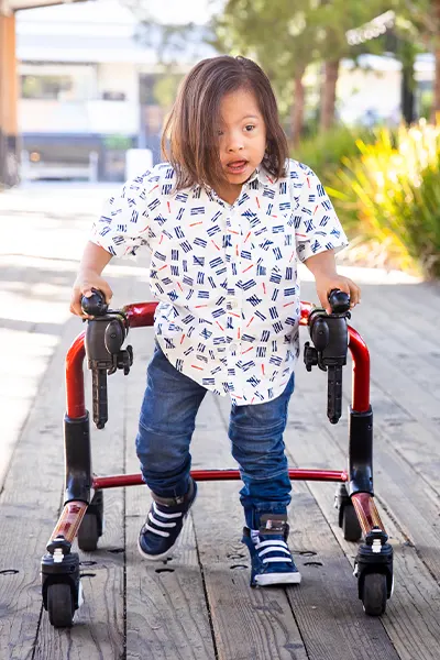 Kind mit Down-Syndrom geht mit einem Posterior-Walker auf einem Holzweg im Freien, unterstützt die Mobilität und Selbstständigkeit.