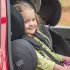 Fröhliches Mädchen sitzt in einem schwarzen Kindersitz im Rücksitz eines roten Autos, hält eine Kuscheldecke und schaut lächelnd zur geöffneten Autotür.