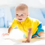 Krabbelndes Baby in gelbem Shirt und blauen Shorts auf hellem Untergrund.