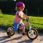 Kind im Alter von 2,5 bis 3 Jahren fährt mit einem rosa Laufrad draußen auf einem Weg, trägt Schutzhelm und lernt spielerisch Gleichgewicht und Mobilität.